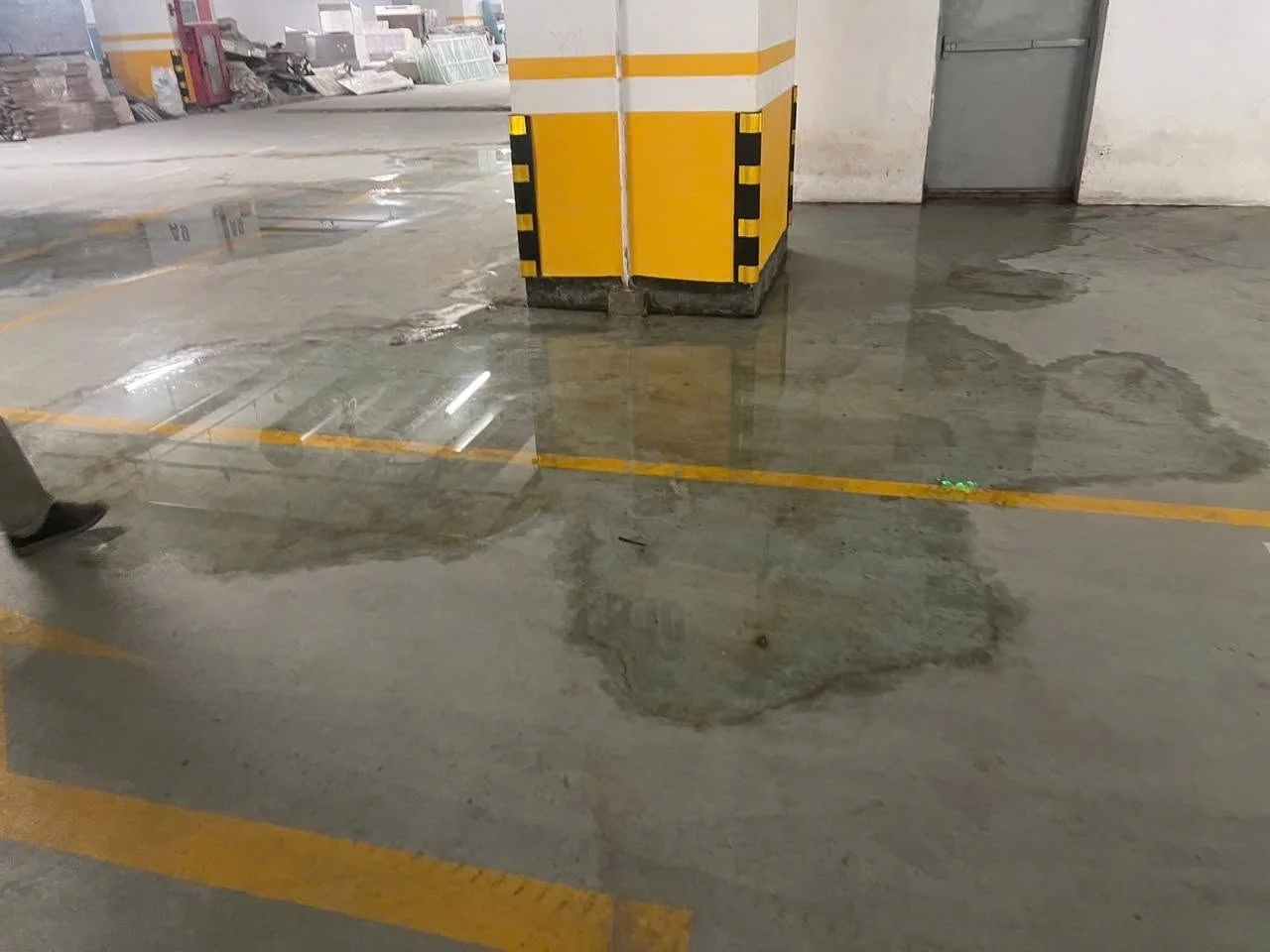Tầng hầm bị thấm nước, sàn bê tông loang lổ, cột trụ vàng trắng, vạch kẻ đường, cửa xám