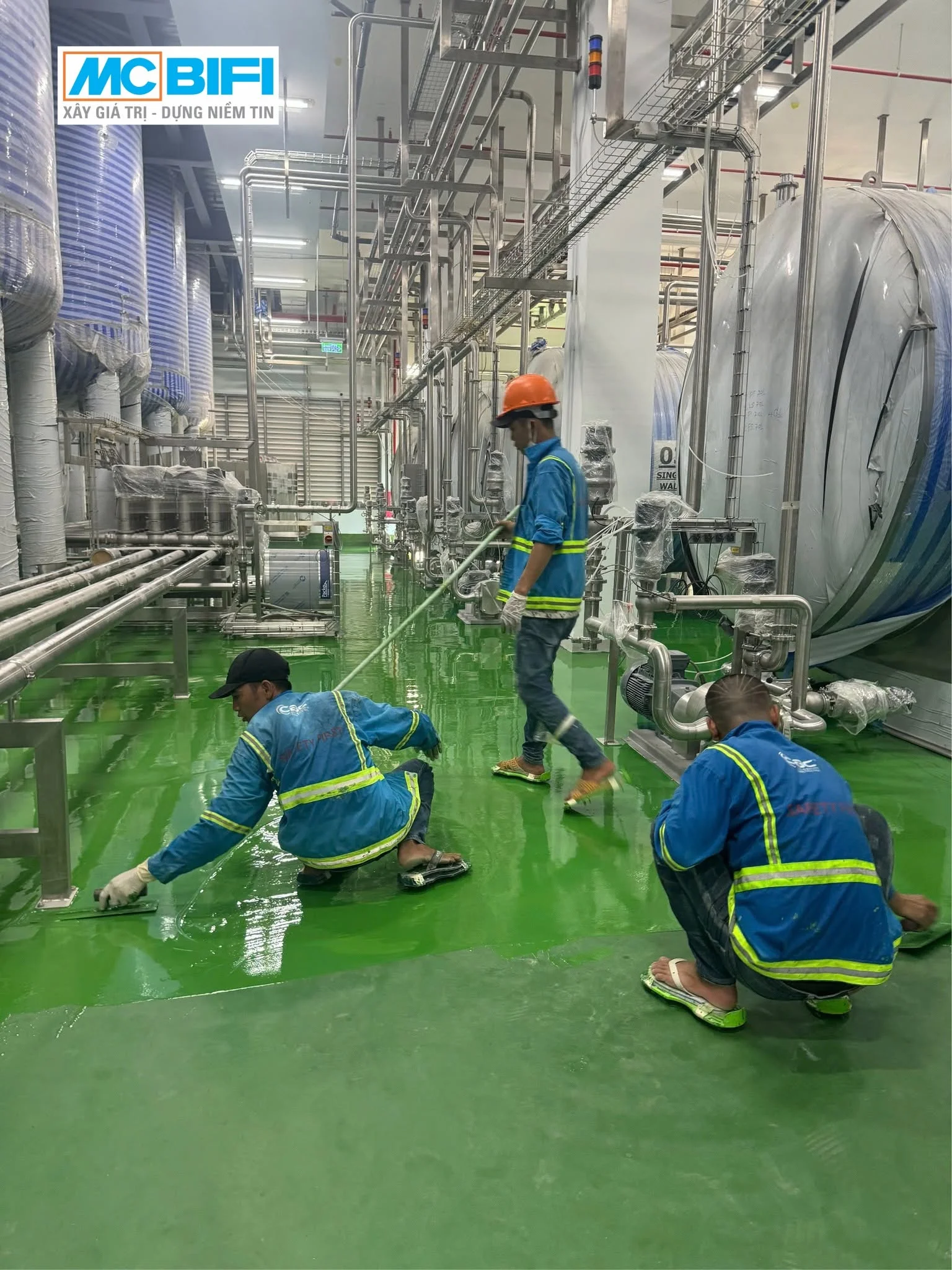 Công nhân thi công sơn epoxy sàn nhà máy công nghiệp với hệ thống ống dẫn và bồn chứa lớn