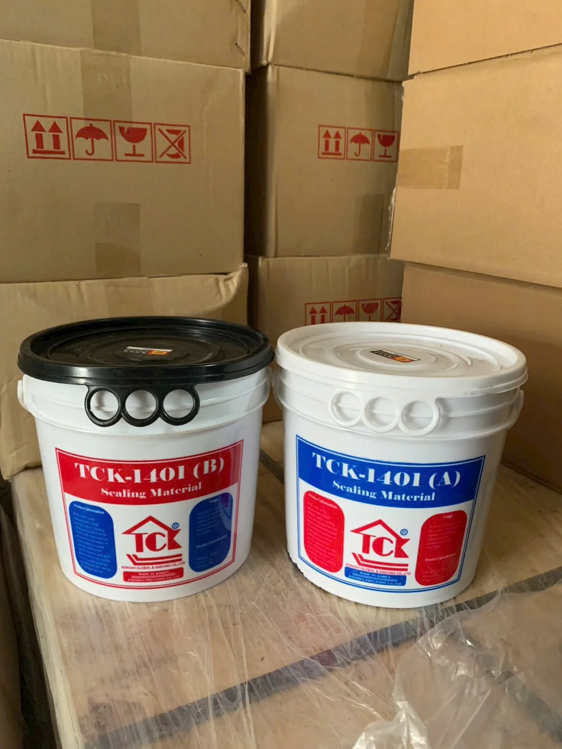 Hai thùng vật liệu trám khe TCK-1401 (A) và (B) màu trắng, có nhãn đỏ xanh, đặt trên nền gỗ, phía sau là thùng carton.